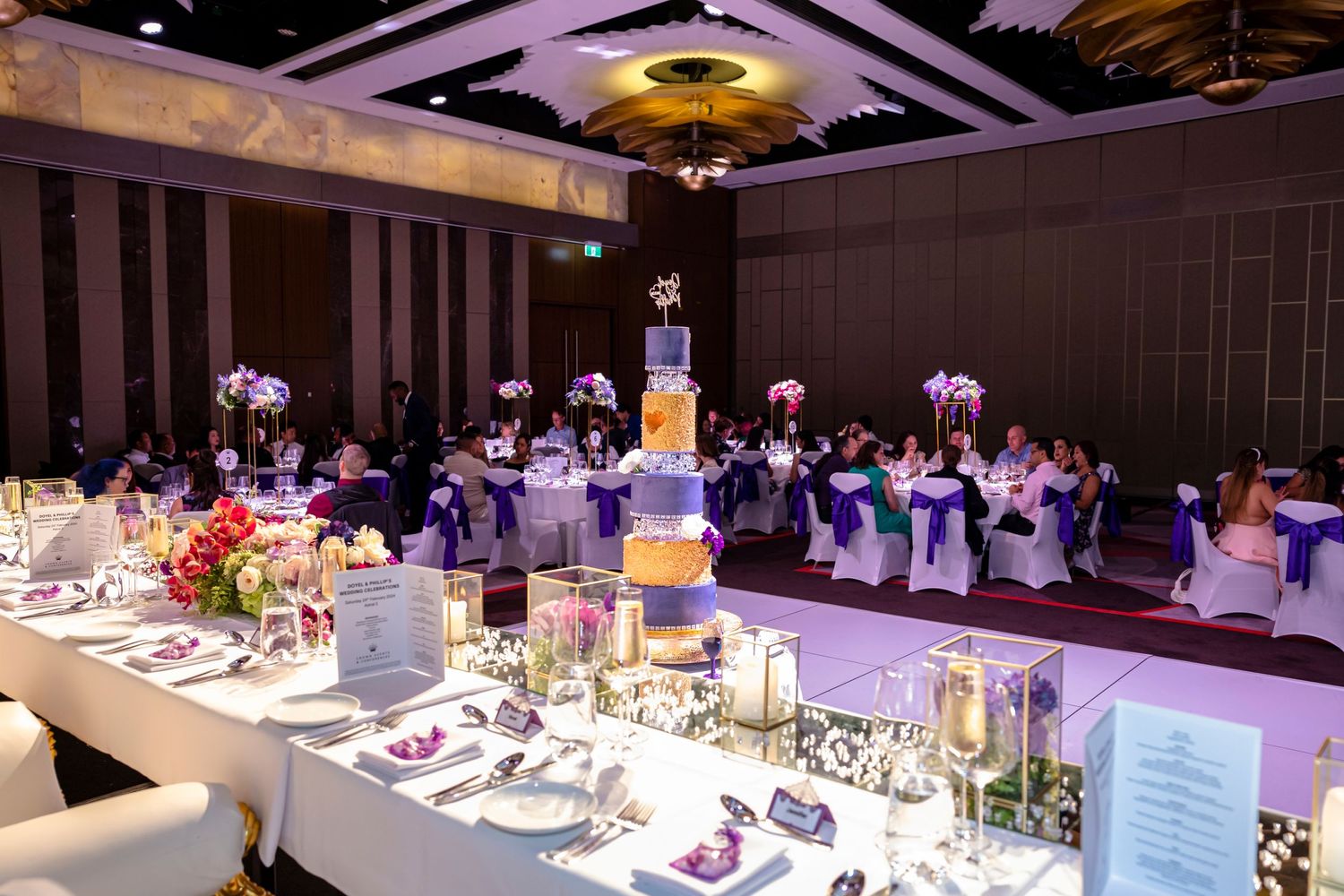 Formal dining tables arranged with white linens and place settings under purple ambient lighting.