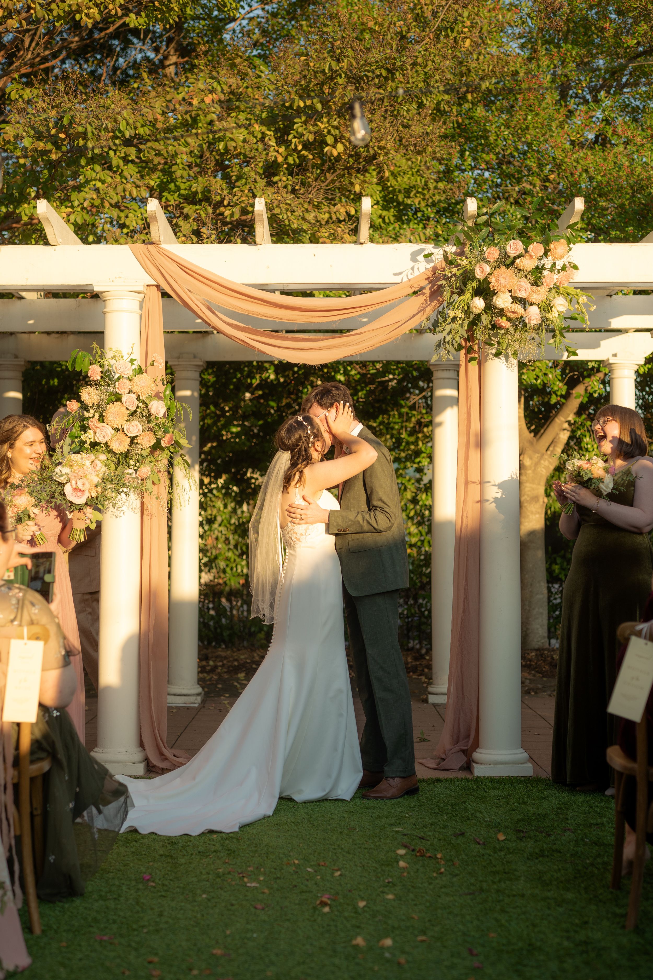Romantic wedding ceremony under white pergola decorated with floral arrangements and fabric draping.