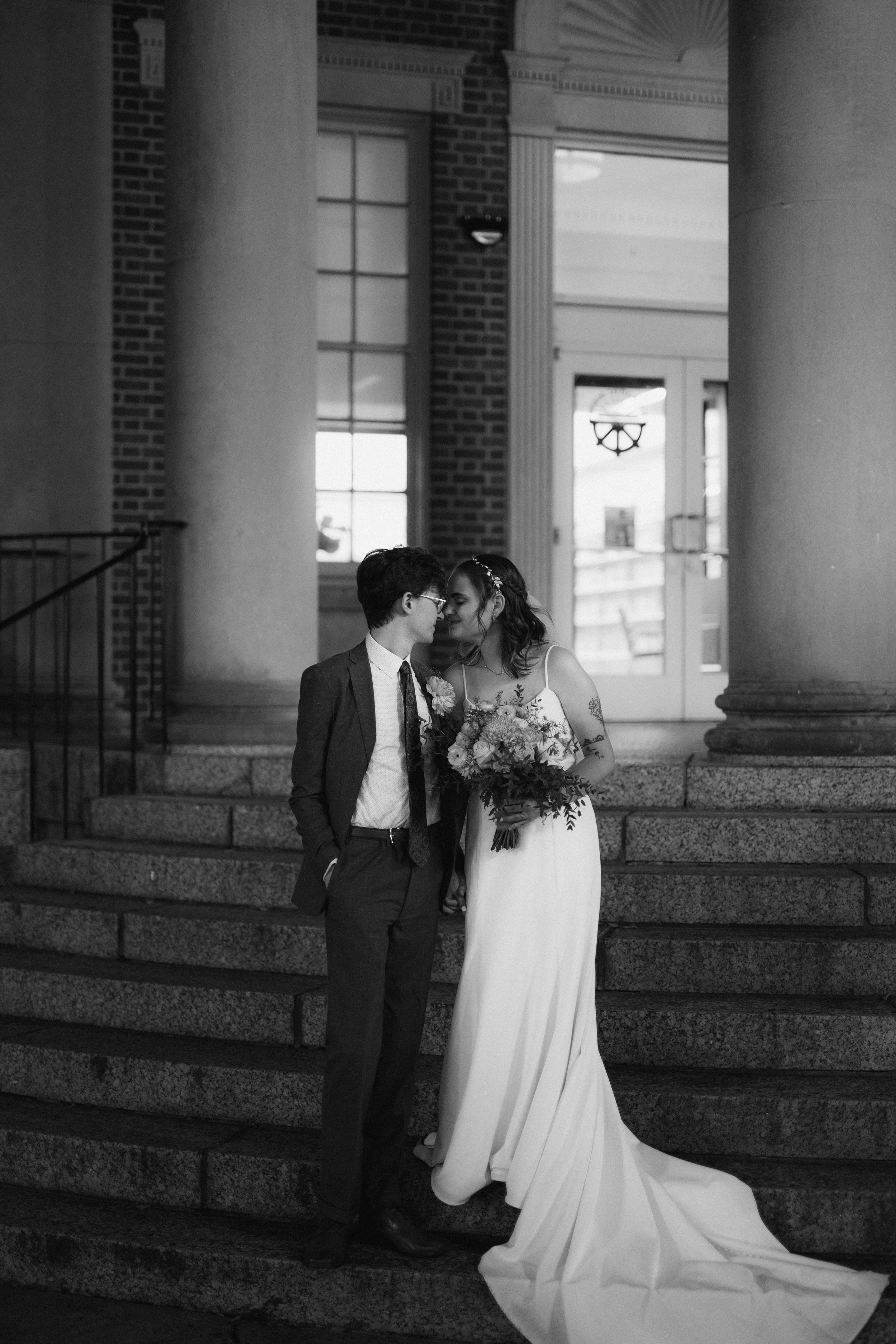 Wedding couple stands on brick steps outside a historic building in black and white.