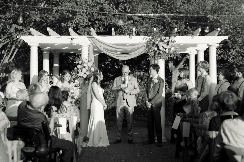 Black and white photo of outdoor wedding ceremony under white pergola with fabric draping.