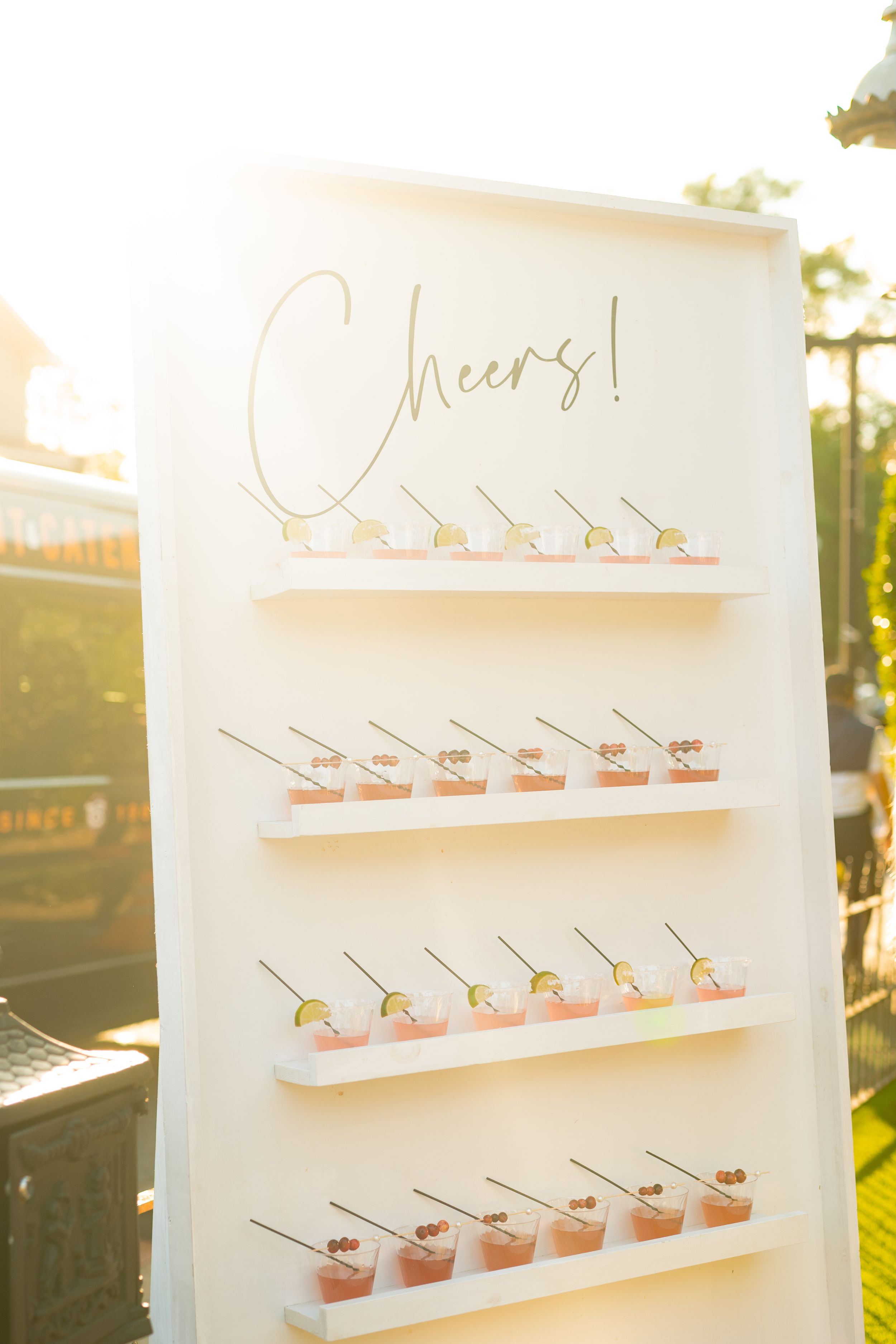 Elegant wedding drink display with white shelves showcasing pink cocktails and gold 'Cheers' text at sunset.