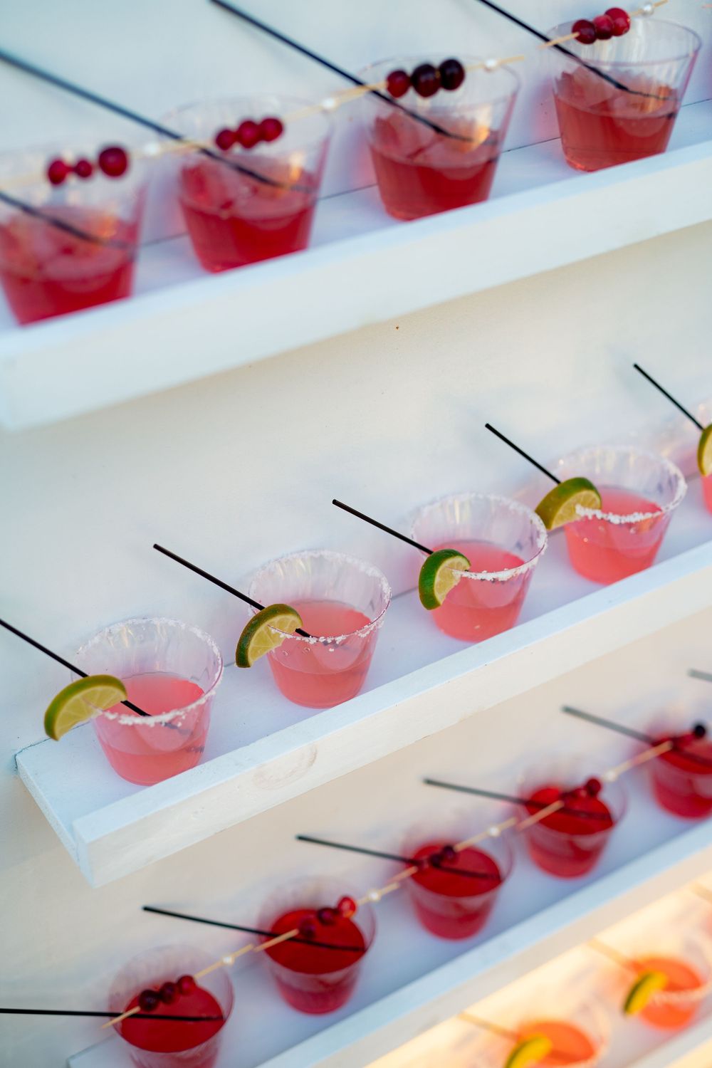 Pink cocktails garnished with cranberries and lime slices arranged on white display shelves.