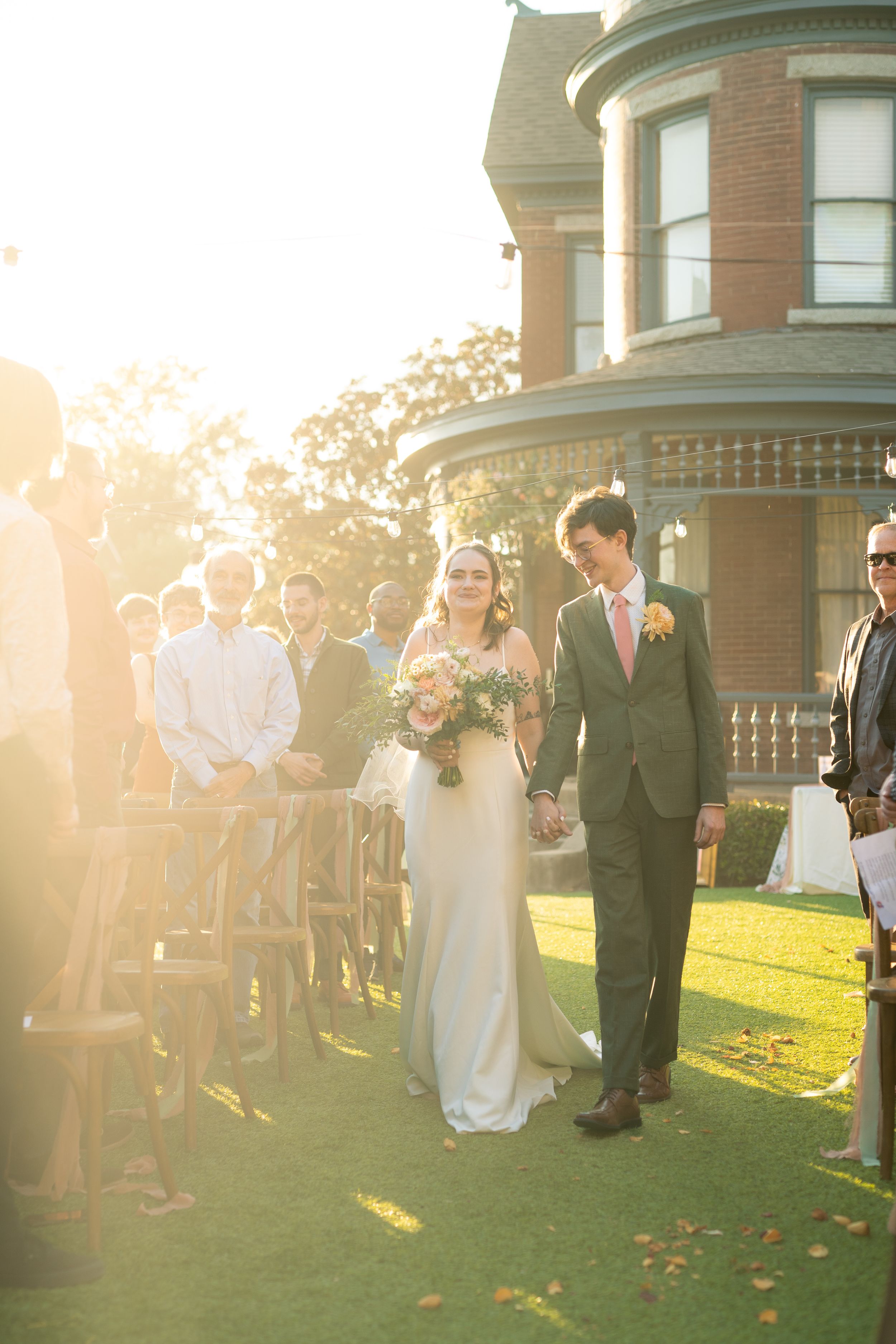 Outdoor wedding ceremony with couple walking on grass against historic brick building in golden light.