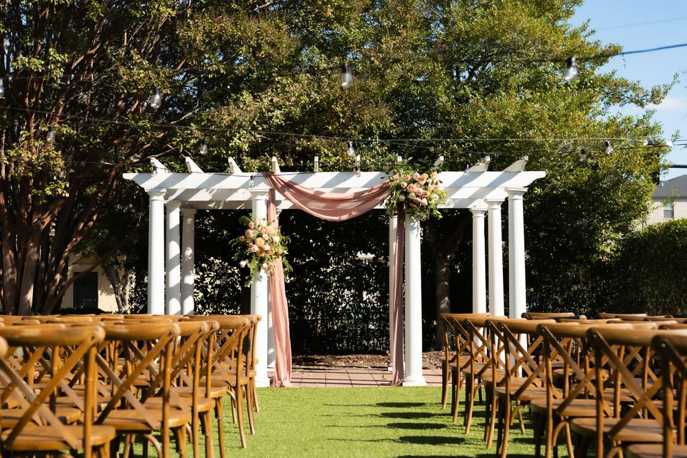 Outdoor wedding ceremony setup with white pergola and wooden cross-back chairs on a grass lawn.