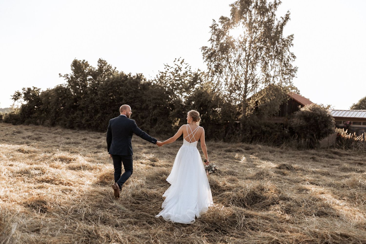 A couple walks through a sunlit field in wedding attire, captured from behind.