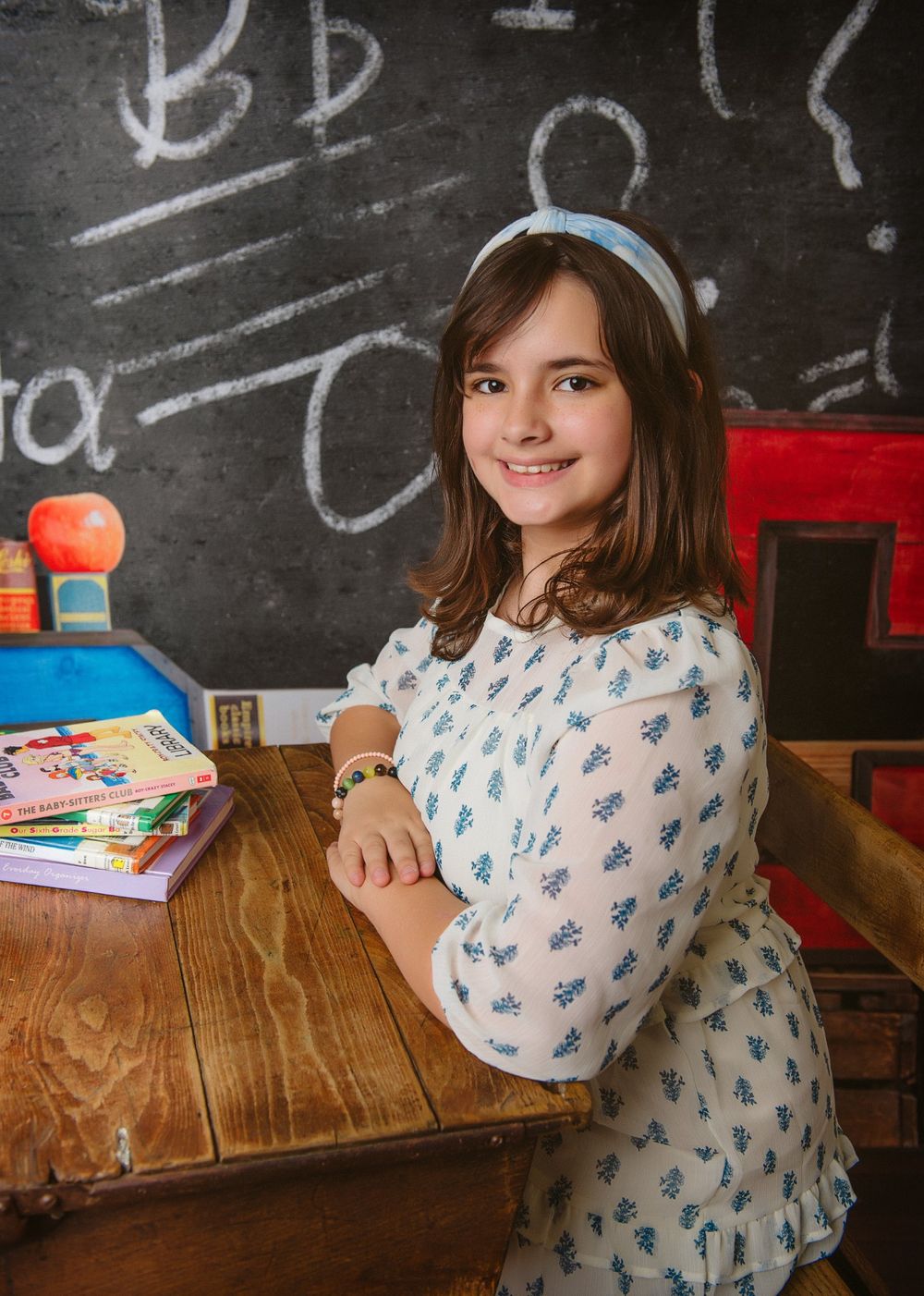 Student sitting at wooden desk with chalkboard background in classroom setting.