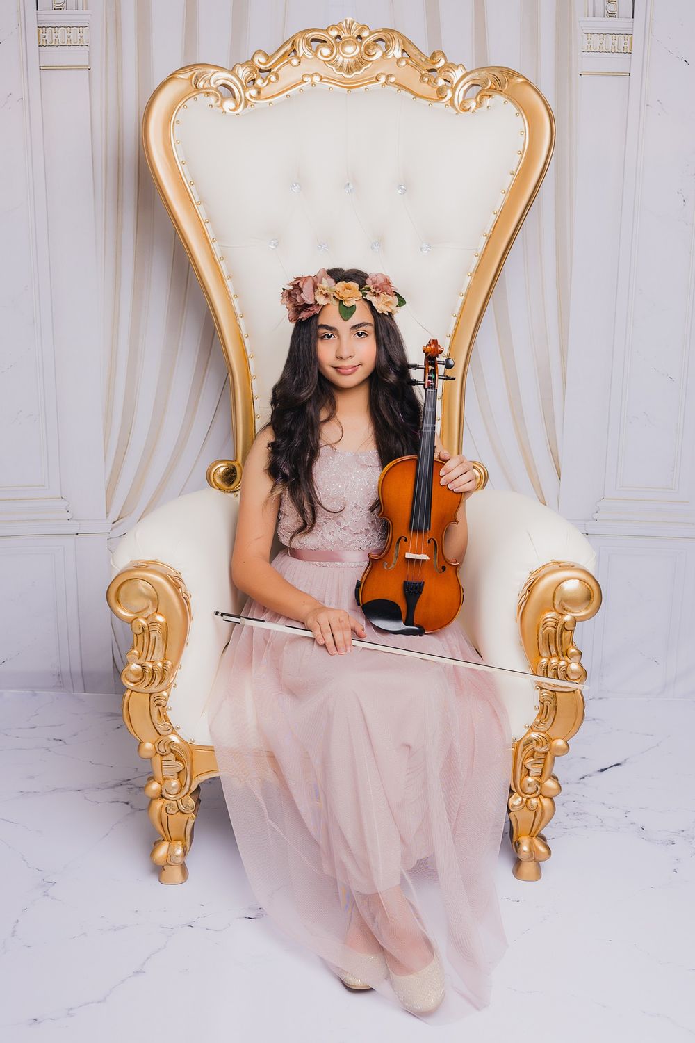 Musician in flowing pink dress and floral crown poses with violin in ornate white throne.