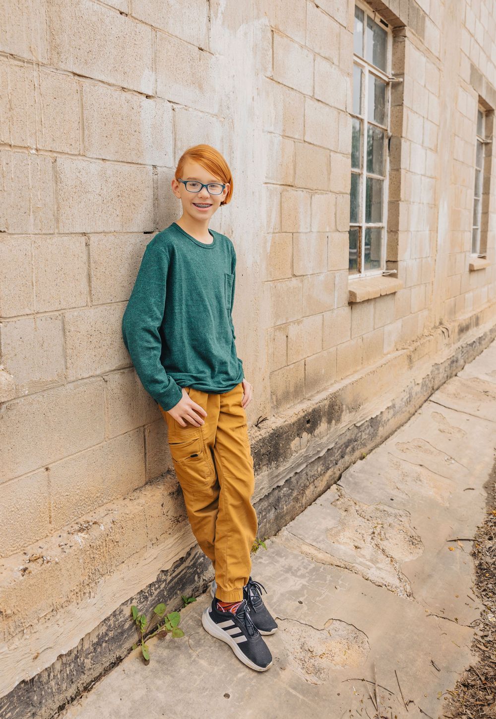 Person wearing green sweater and yellow pants leans against stone wall near windows.