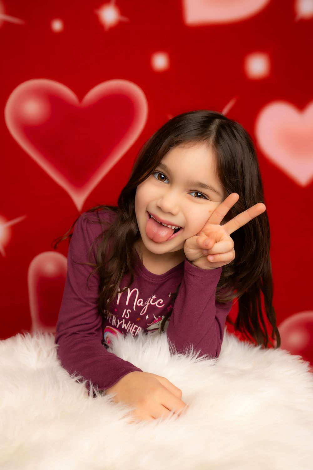 Child laying on white fur with heart bokeh background showing peace sign in purple shirt.