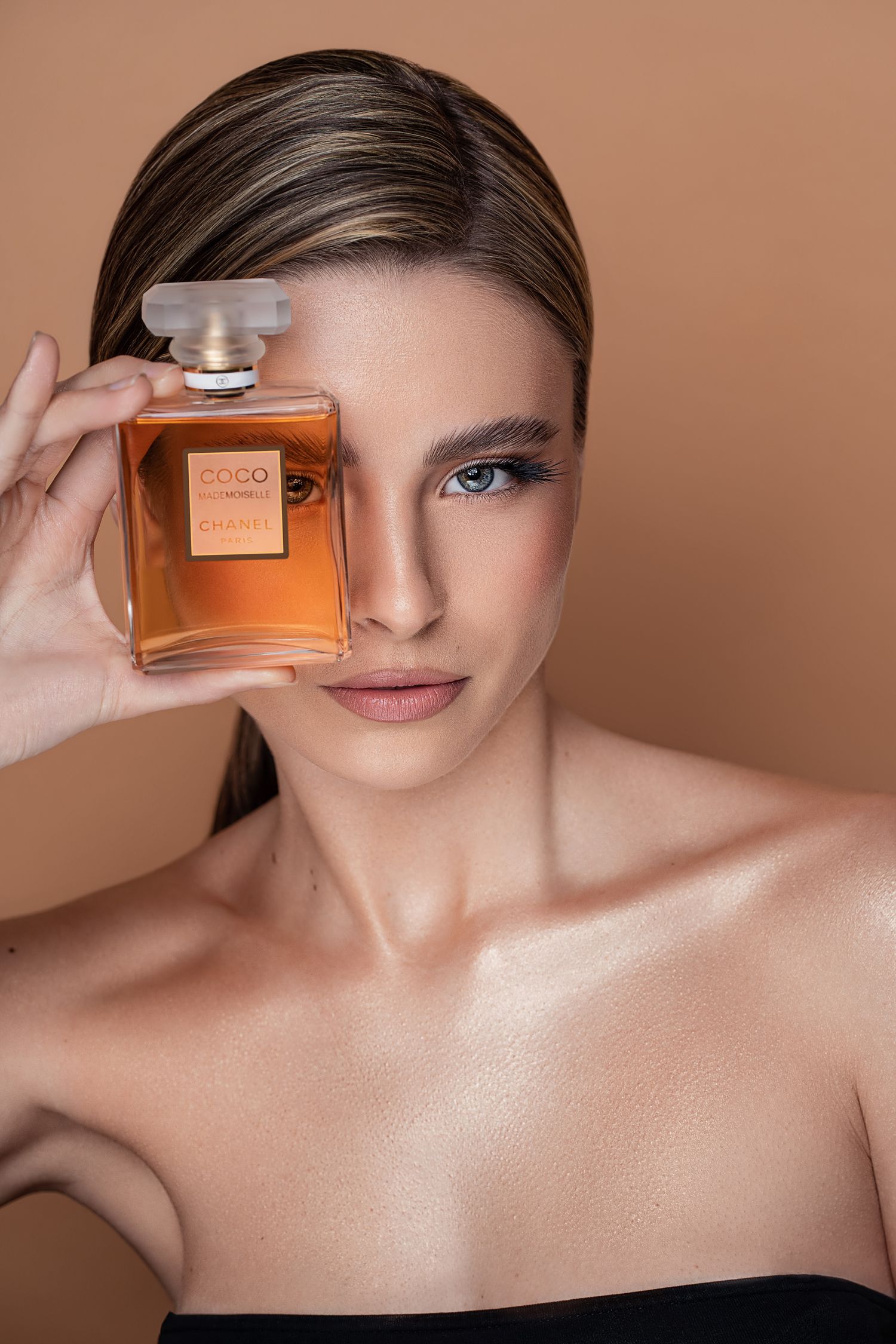 A bottle of Coco perfume held next to bare shoulders against a warm brown background in a beauty product advertisement.