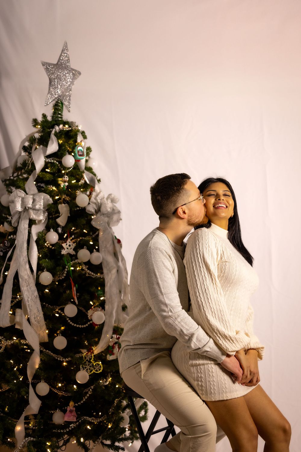 Couple in winter white outfits posing romantically by decorated Christmas tree.