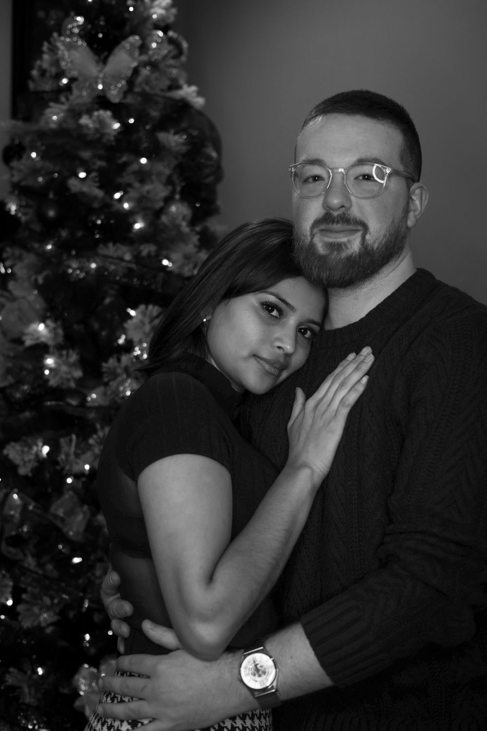 A tender black and white portrait of a couple posing by their decorated Christmas tree.