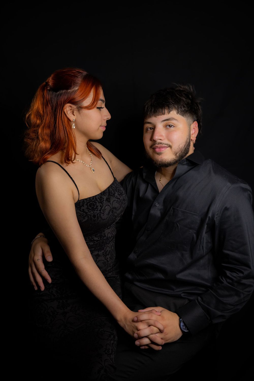 Studio portrait of a couple in black attire holding hands against dark background.