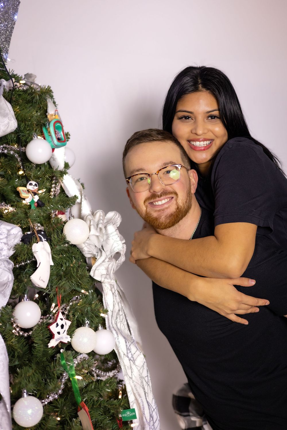 Couple embraces and smiles while posing next to an elegantly decorated Christmas tree with white ornaments.