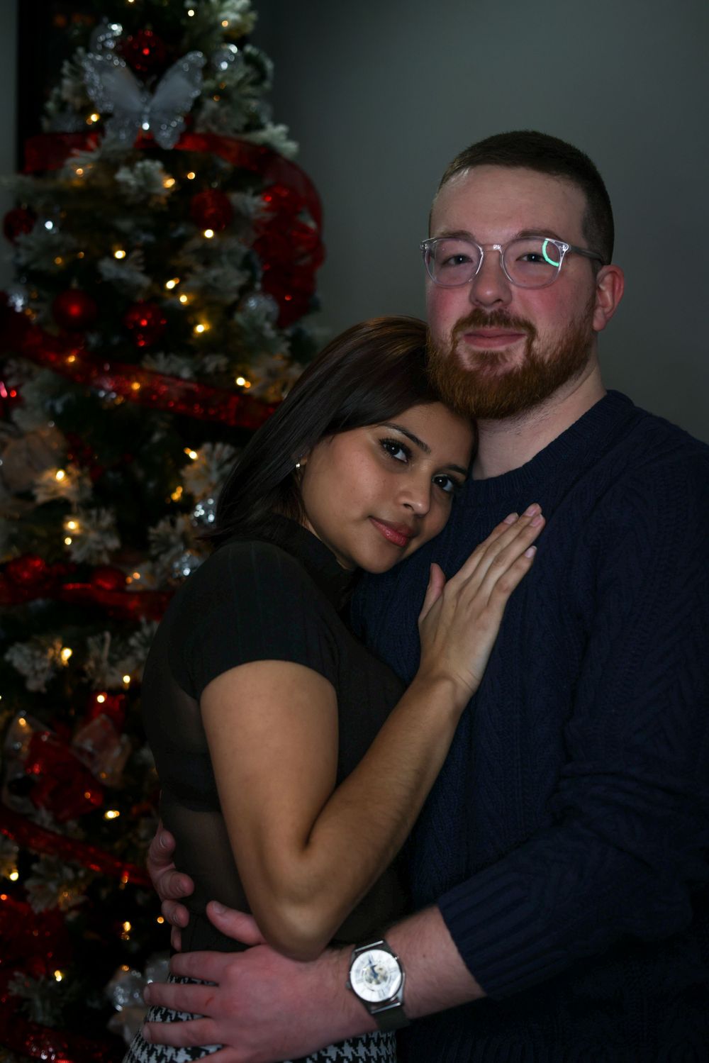 A couple poses for an intimate portrait in front of their Christmas tree with red decorations.