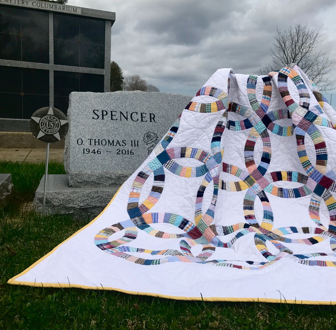 A double wedding ring memory quilt lays over a grey head stone on a cloudy day.