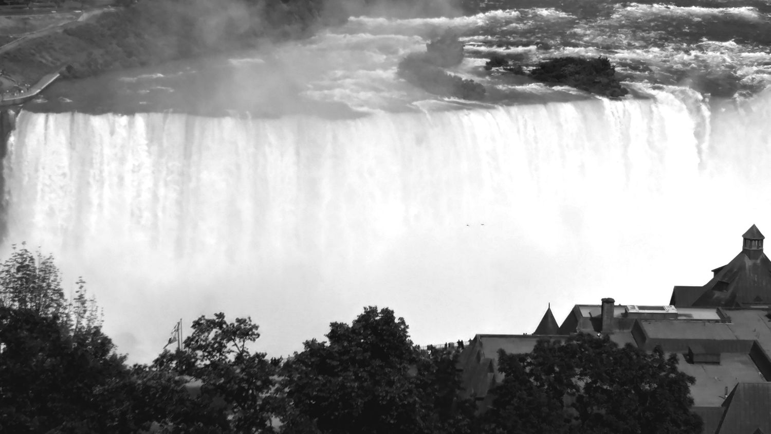 A silhouetted skyline of buildings with Niagara Falls cascading in the misty background, captured in black and white.
