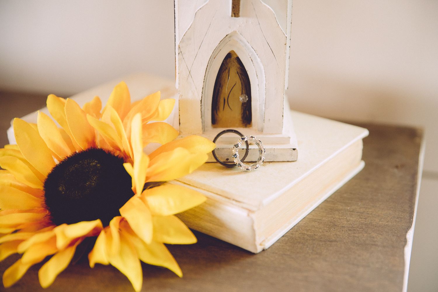 Wedding rings displayed with a decorative white arch and sunflower on a white book.