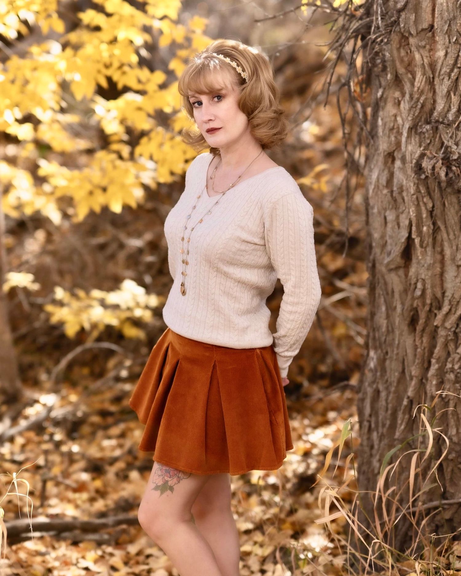 A vintage-inspired fall portrait featuring a cream cardigan and rust-colored corduroy skirt against autumn foliage.