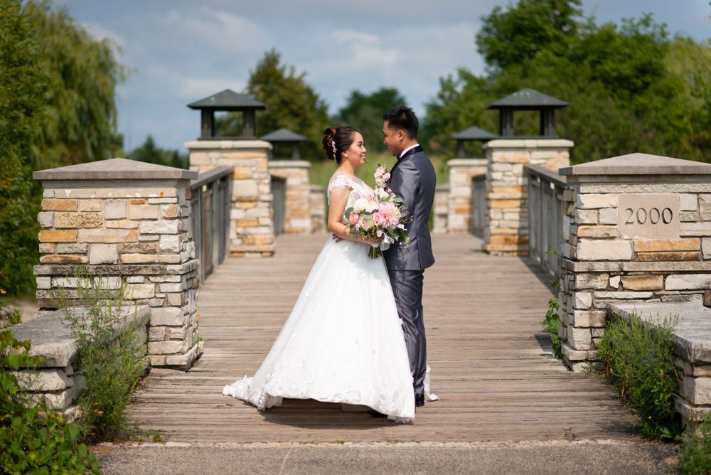 A wedding couple stands on a stone and wooden bridge surrounded by lush greenery on a sunny day.