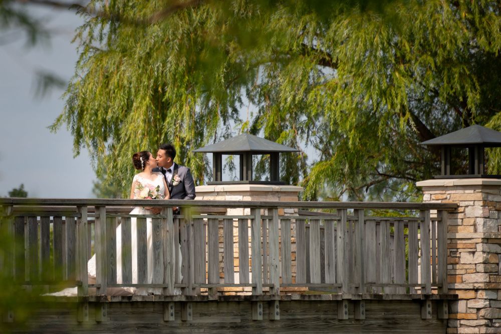 Newlyweds share a romantic moment on a wooden bridge with willow trees in the background.
