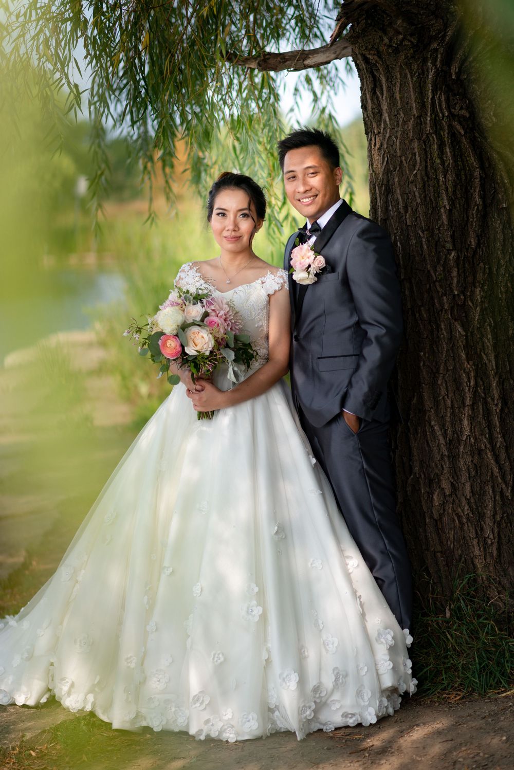A wedding couple poses under a weeping willow tree with a beautiful floral bouquet.
