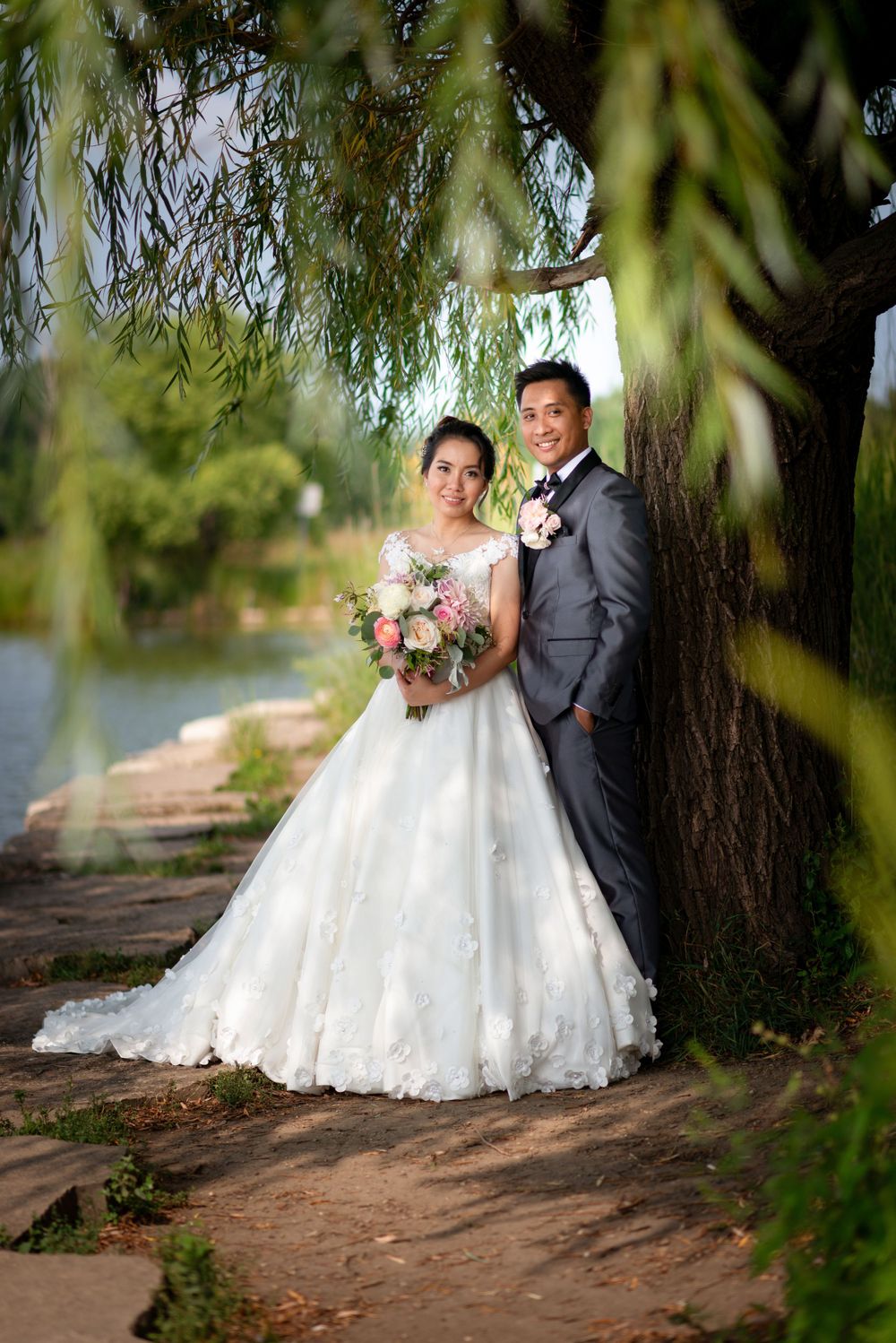 A couple in wedding attire poses beneath drooping willow branches by a waterfront.