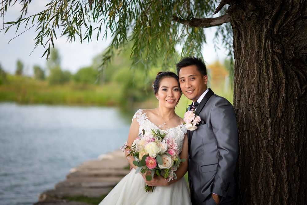 A couple in wedding attire poses by a lake under a willow tree with pink and white flowers.