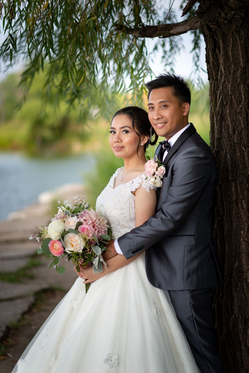 A wedding couple stands against a willow tree holding a pink and white floral bouquet.