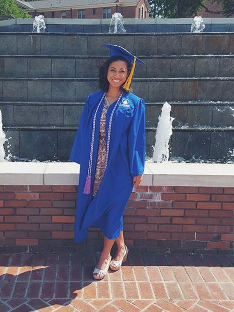 A graduation photo showing someone in blue cap and gown posing against a brick wall.