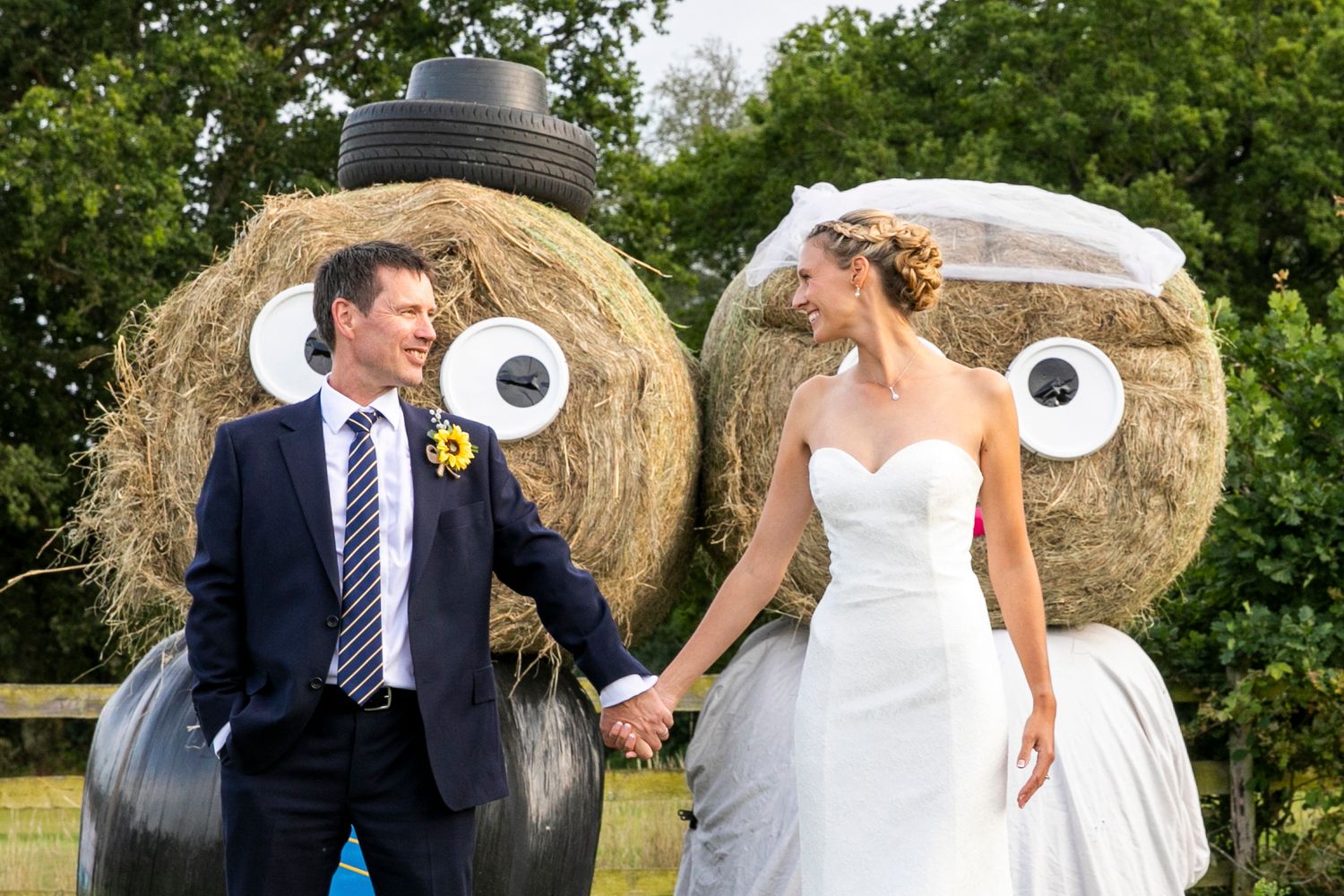 Wedding couple poses playfully near rustic hay bales in a countryside setting.