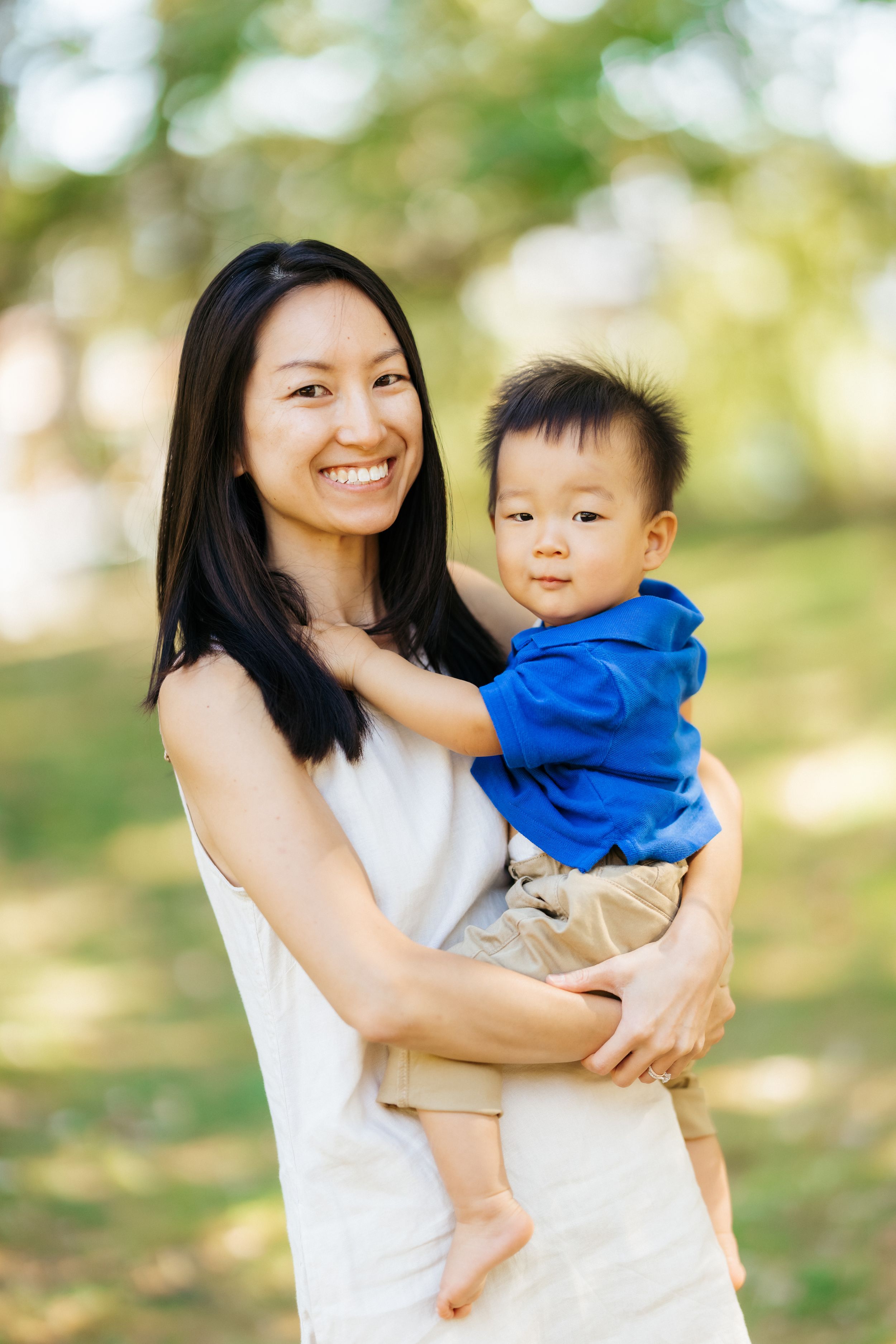 A mother holds her young child while standing in a sunny park setting.