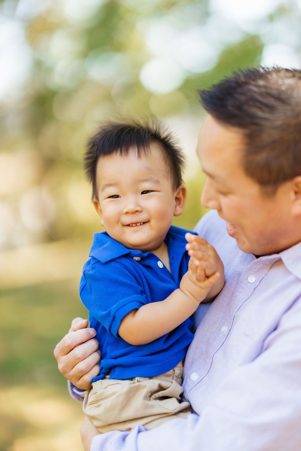 A joyful toddler in a blue shirt smiles while being held outdoors in a natural setting.