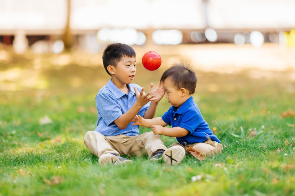 Two children play with a red ball while sitting on grass in a park.