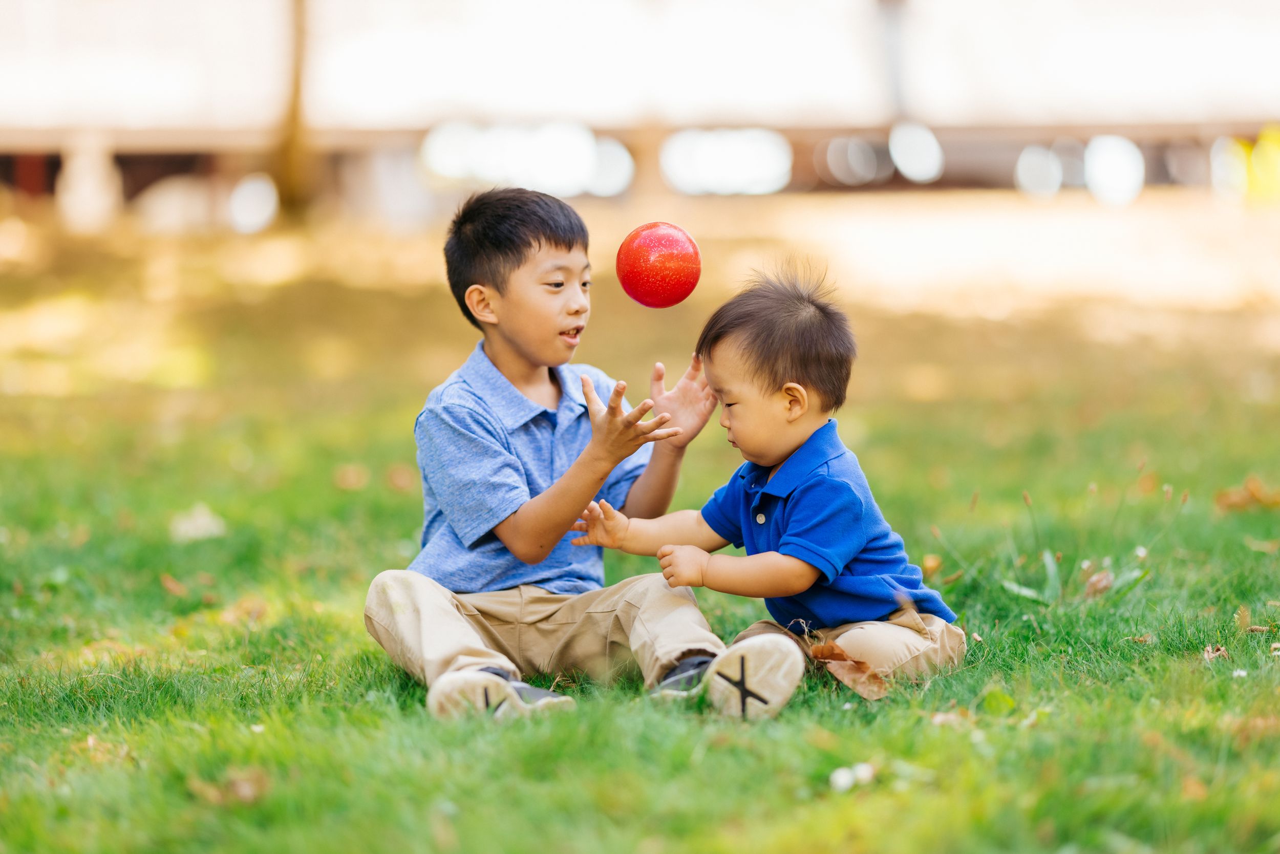 Two children play with a red ball while sitting on grass in a park.