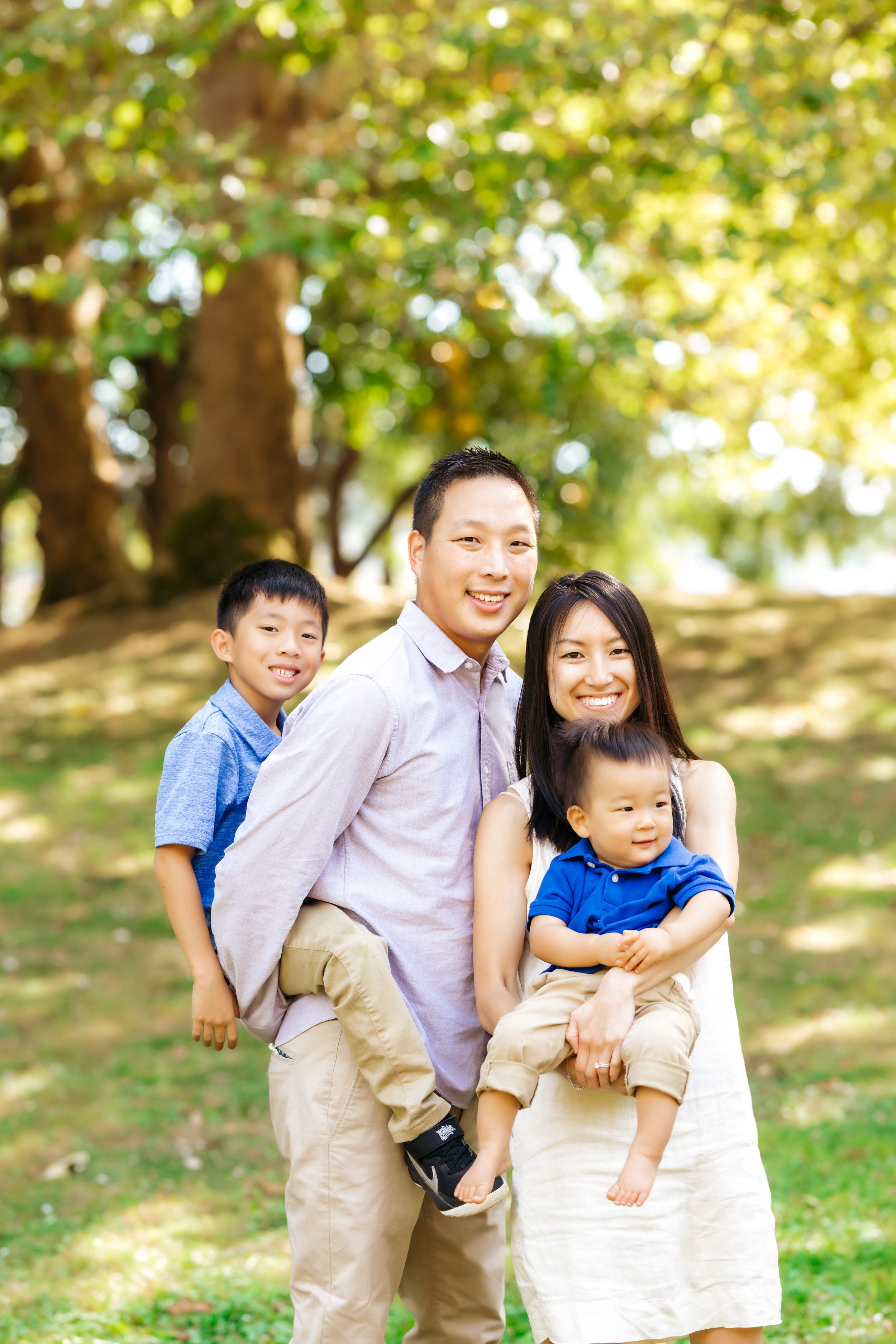 A family of four poses together outdoors on a sunny day in a park.