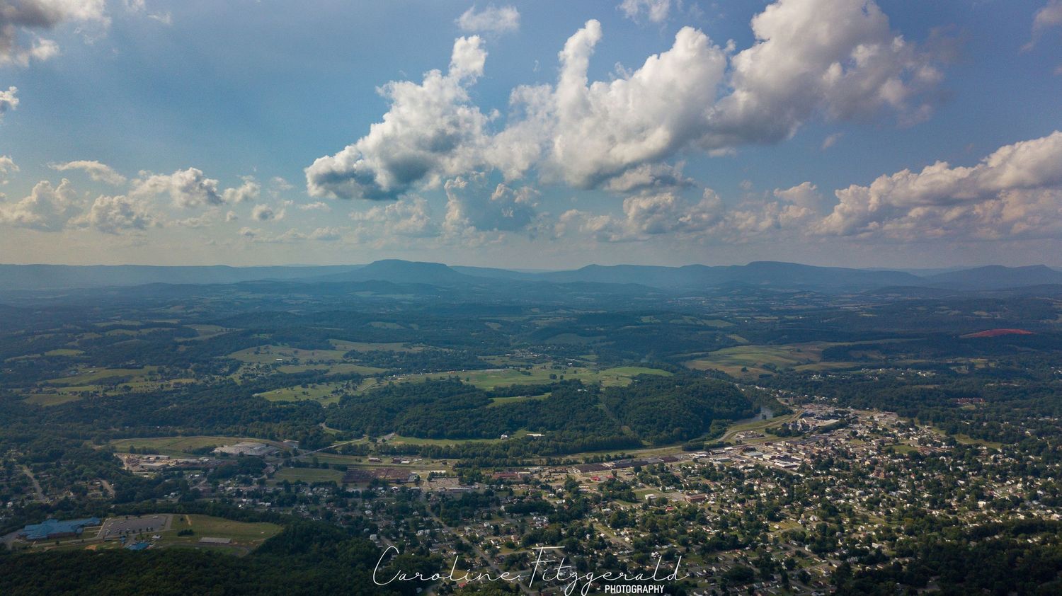 Aerial view of a mountain valley with puffy white clouds and blue skies over green terrain.