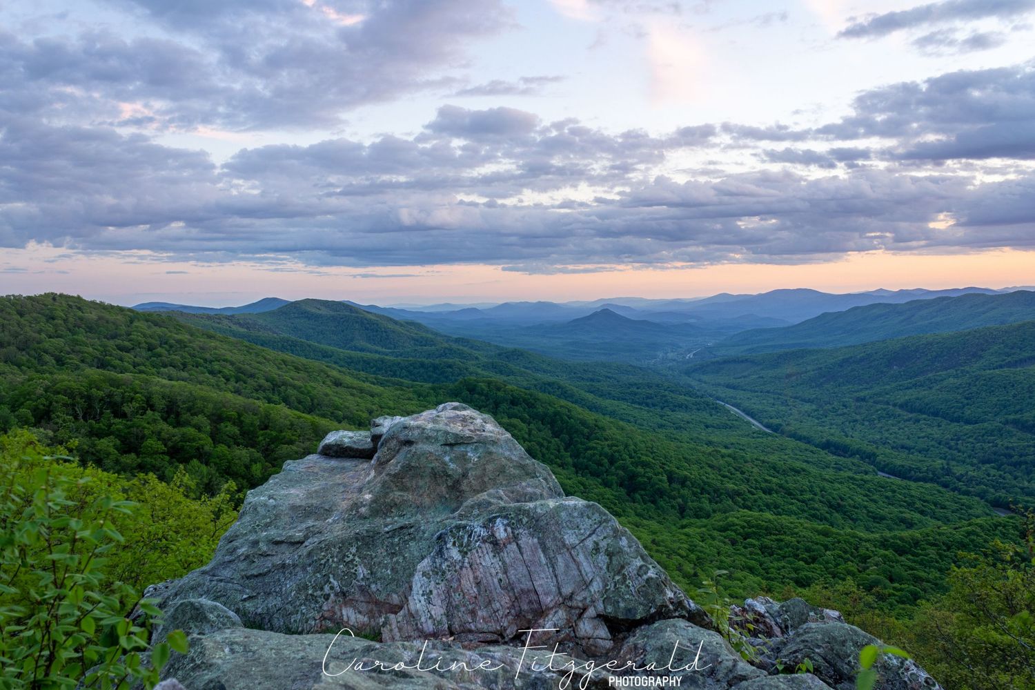 Rocky mountain overlook with dramatic cloudy sky and rolling green hills stretching to the horizon.