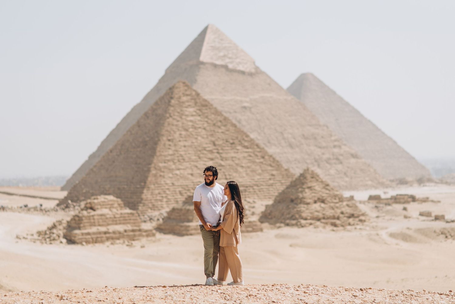 A couple stands together in front of the ancient pyramids of Giza in Egypt on a hazy desert day.