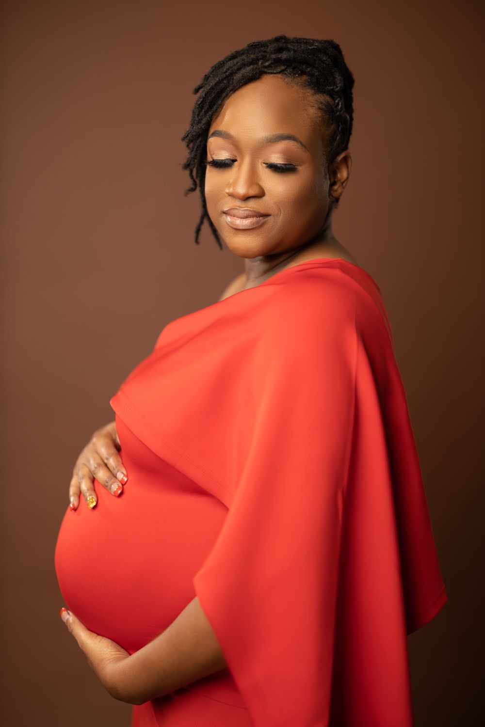 Maternity portrait of expectant mother in flowing red dress against brown background.