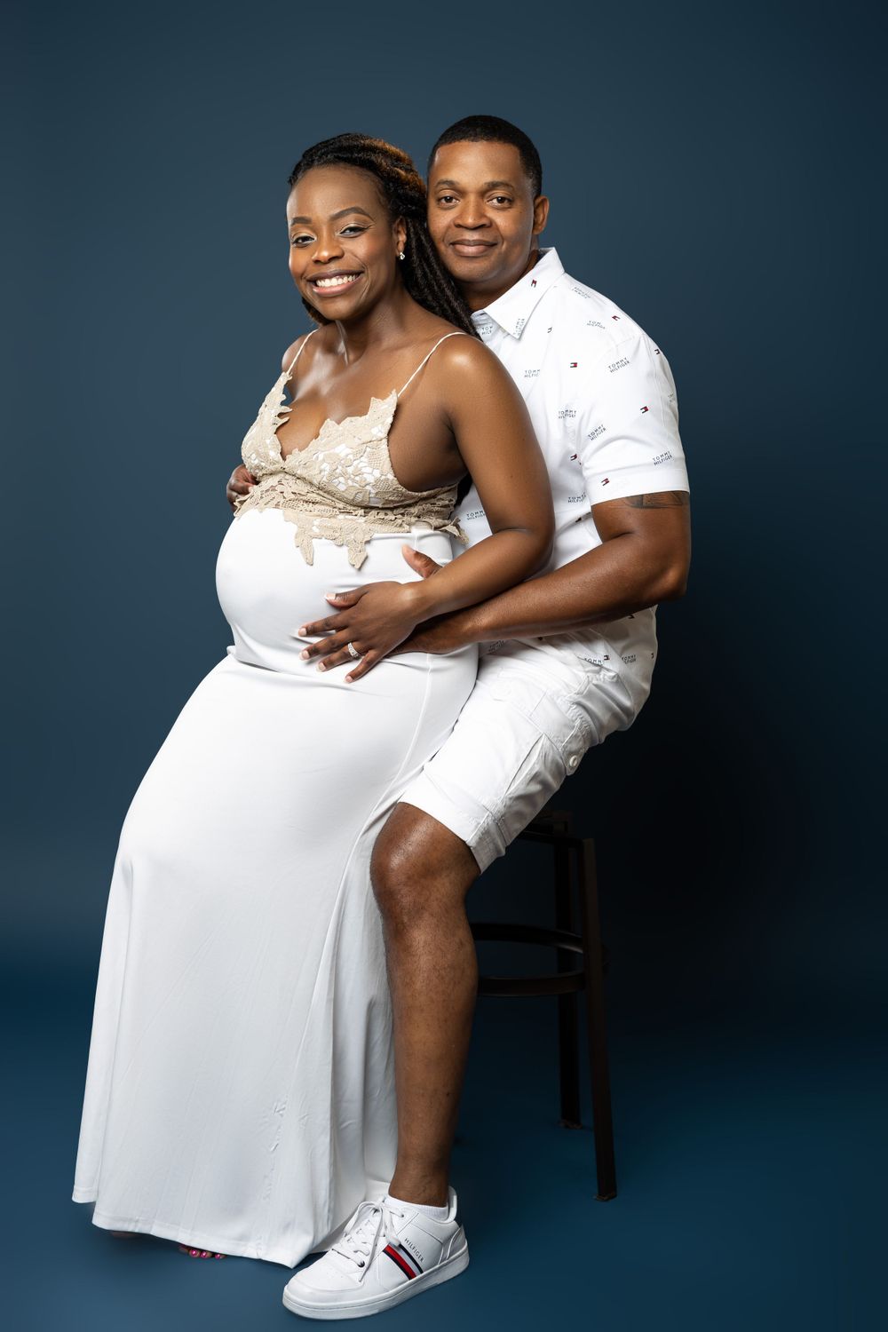 Maternity portrait of an expecting couple in white outfits against dark blue background.