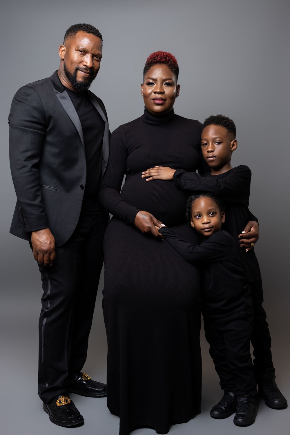Family maternity portrait in matching black outfits against grey background.