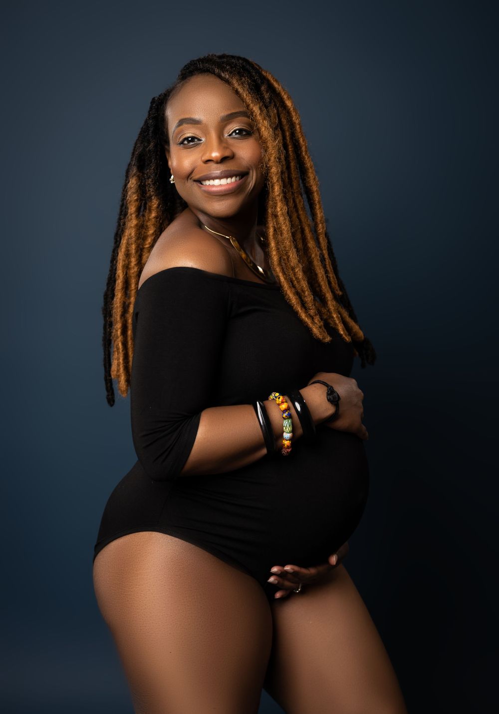 Maternity portrait of a woman in black bodysuit with golden locs against dark background.