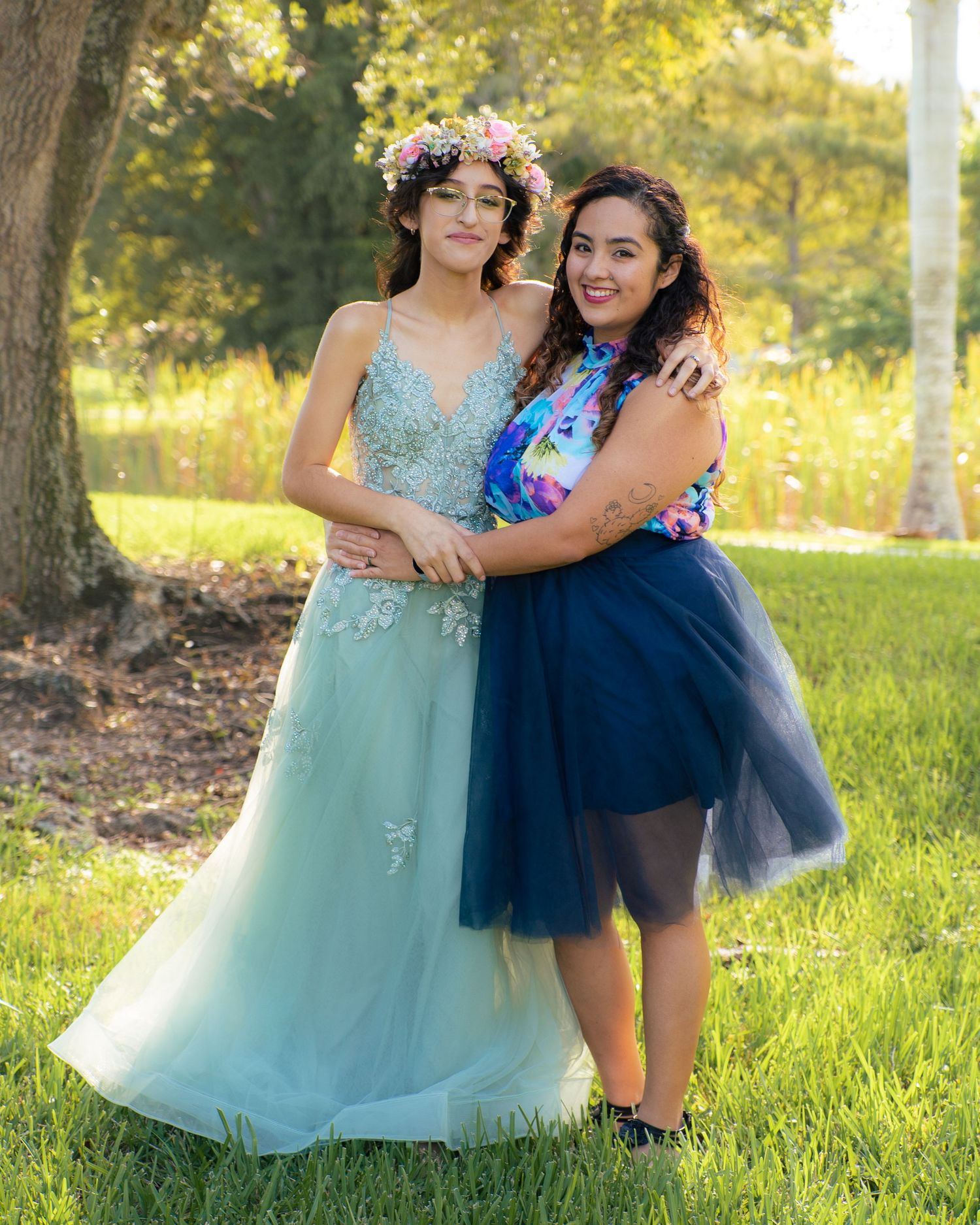 Two young women in elegant dresses pose together outdoors in a lush green park setting.