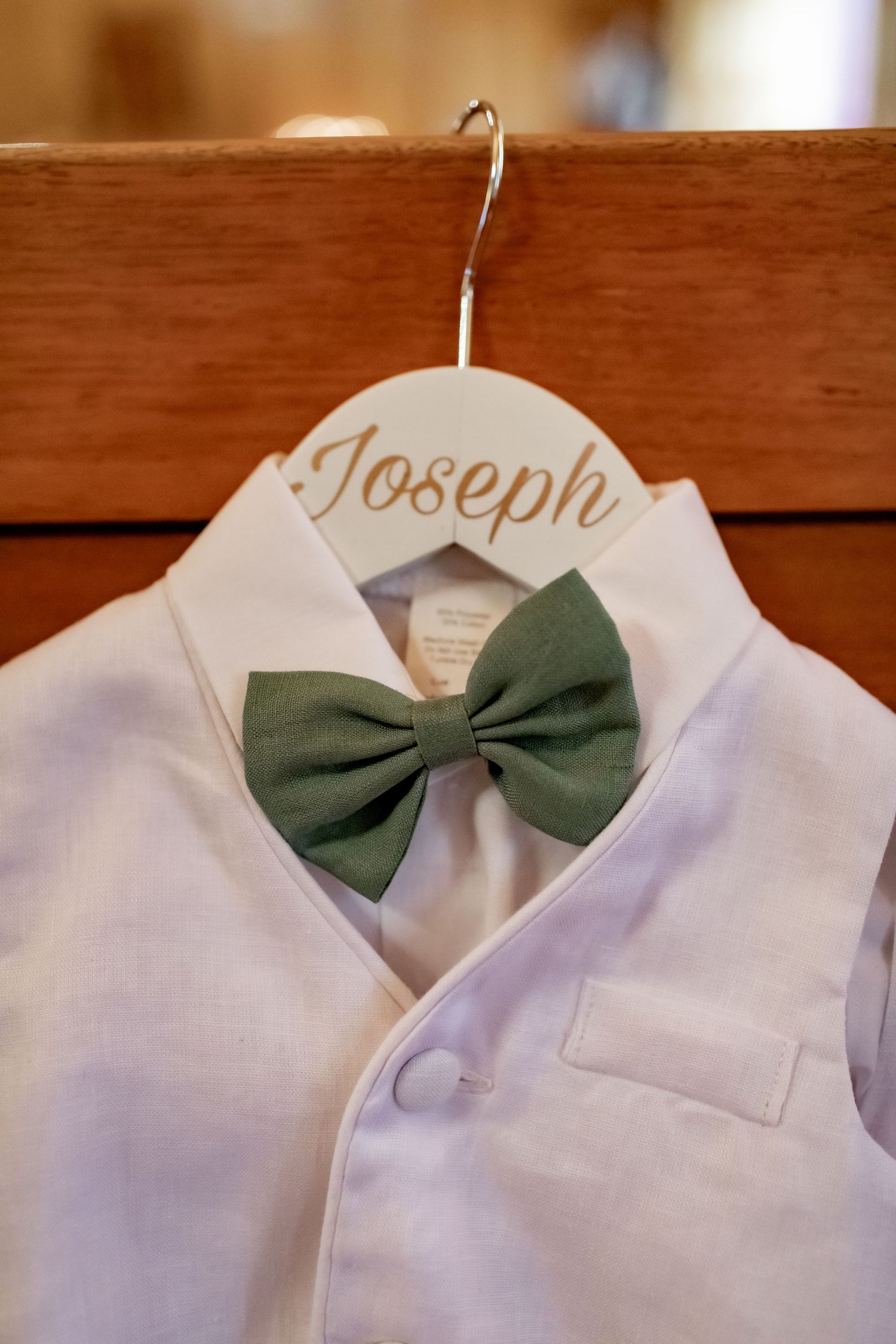 White dress shirt with sage green bowtie hanging on wooden hanger labeled 'Joseph'.