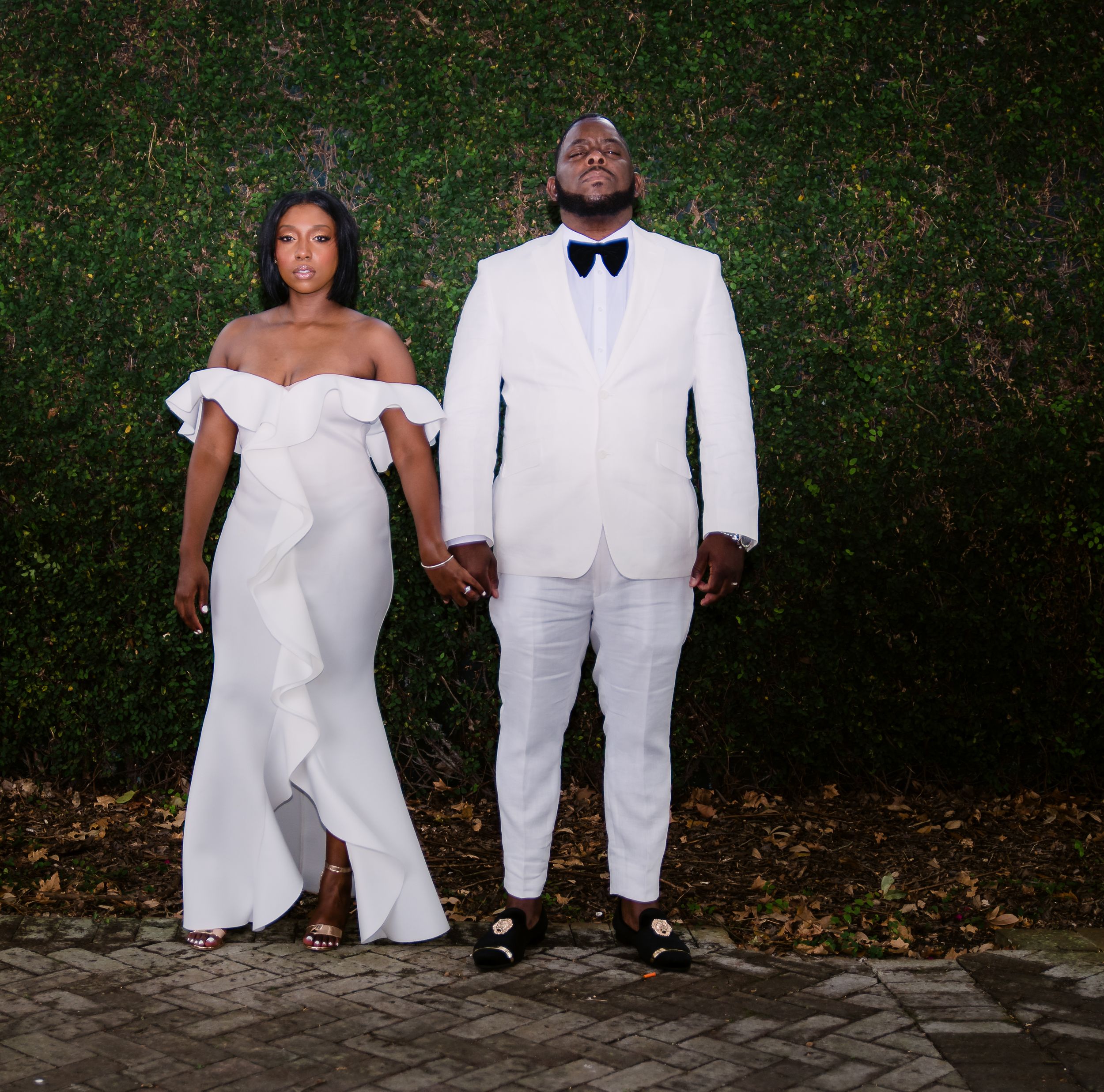 A formal couple in white attire poses against a hedge wall in an artistic wedding photo.