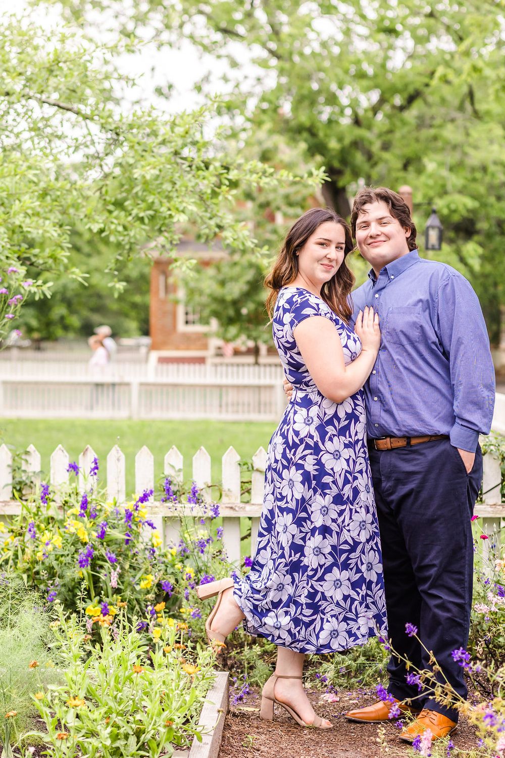 A couple poses in a garden with white picket fence and blooming flowers.