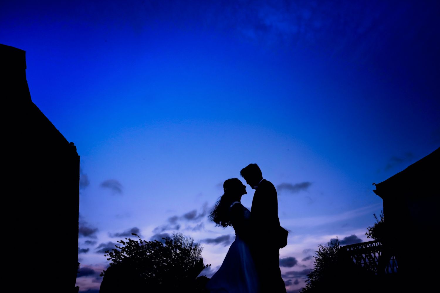 Silhouette of couple embracing against deep blue twilight sky during romantic wedding moment.