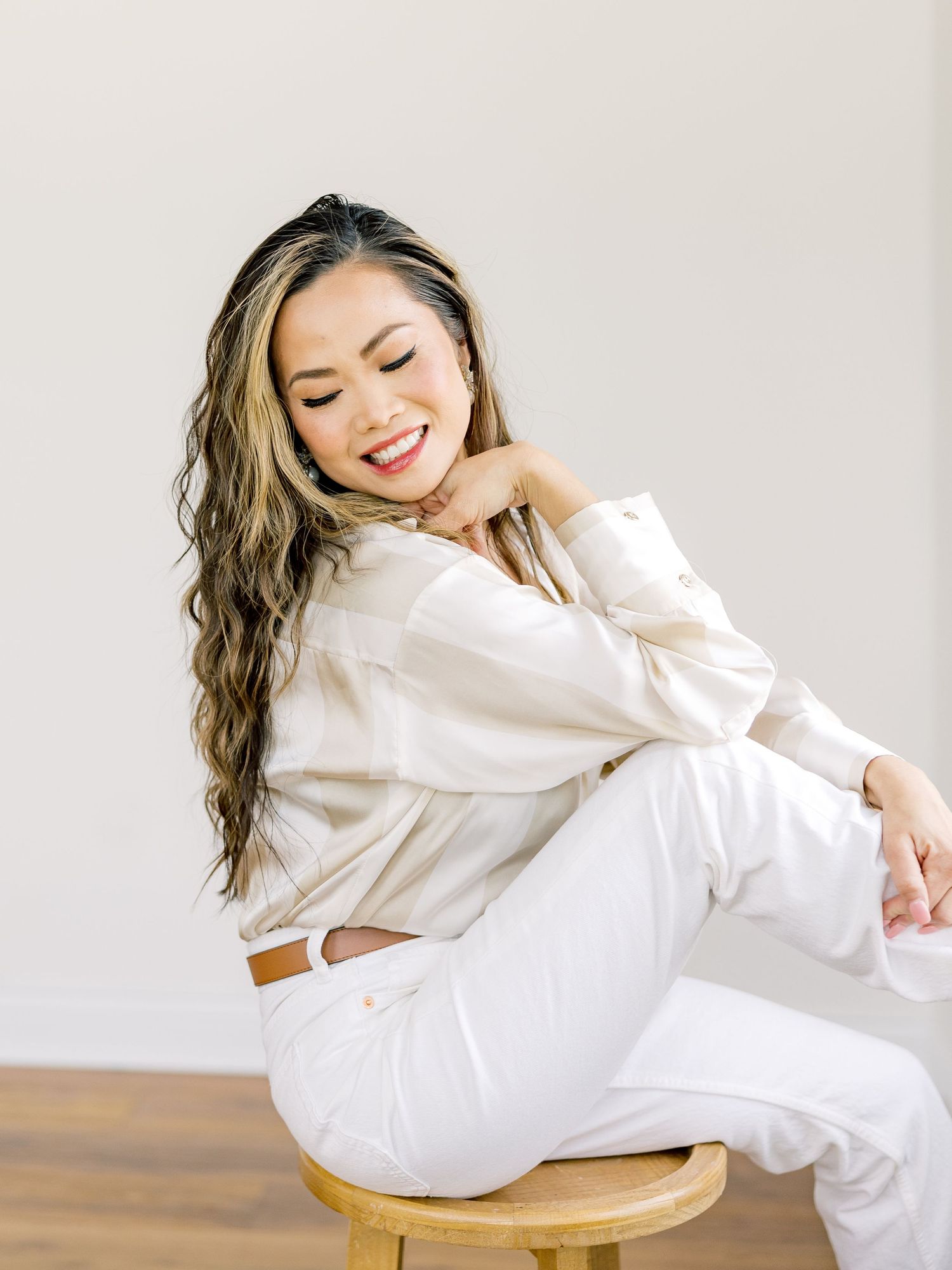 Casual lifestyle portrait in all-white outfit sitting on wooden stool against neutral background.