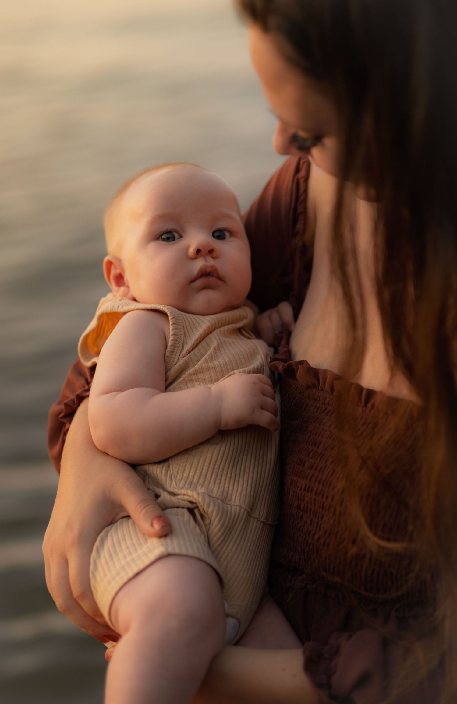 Baby in knit outfit gazes up while being held at sunset by water.